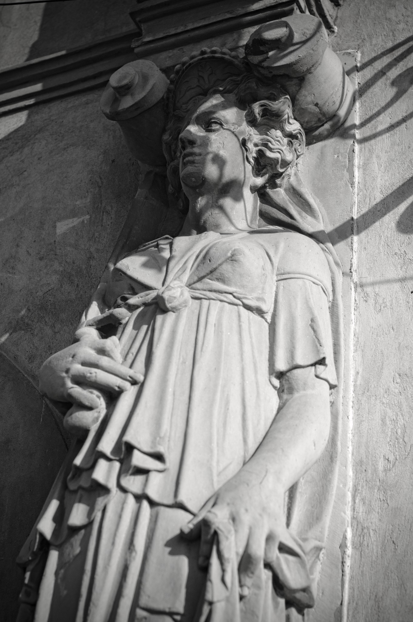 Czarno-białe zdjęcie kamiennej rzeźby kobiety w drapowanej szacie, z dzbanami na głowie, umieszczonej na ścianie budynku, oświetlonej z góry.