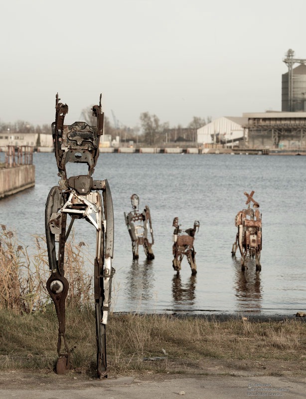 Rzeźby robotów ze złomu, stojące w wodzie, na tle przemysłowego krajobrazu. Pierwszy plan: figura z podniesionymi rękami. Artystyczna instalacja.