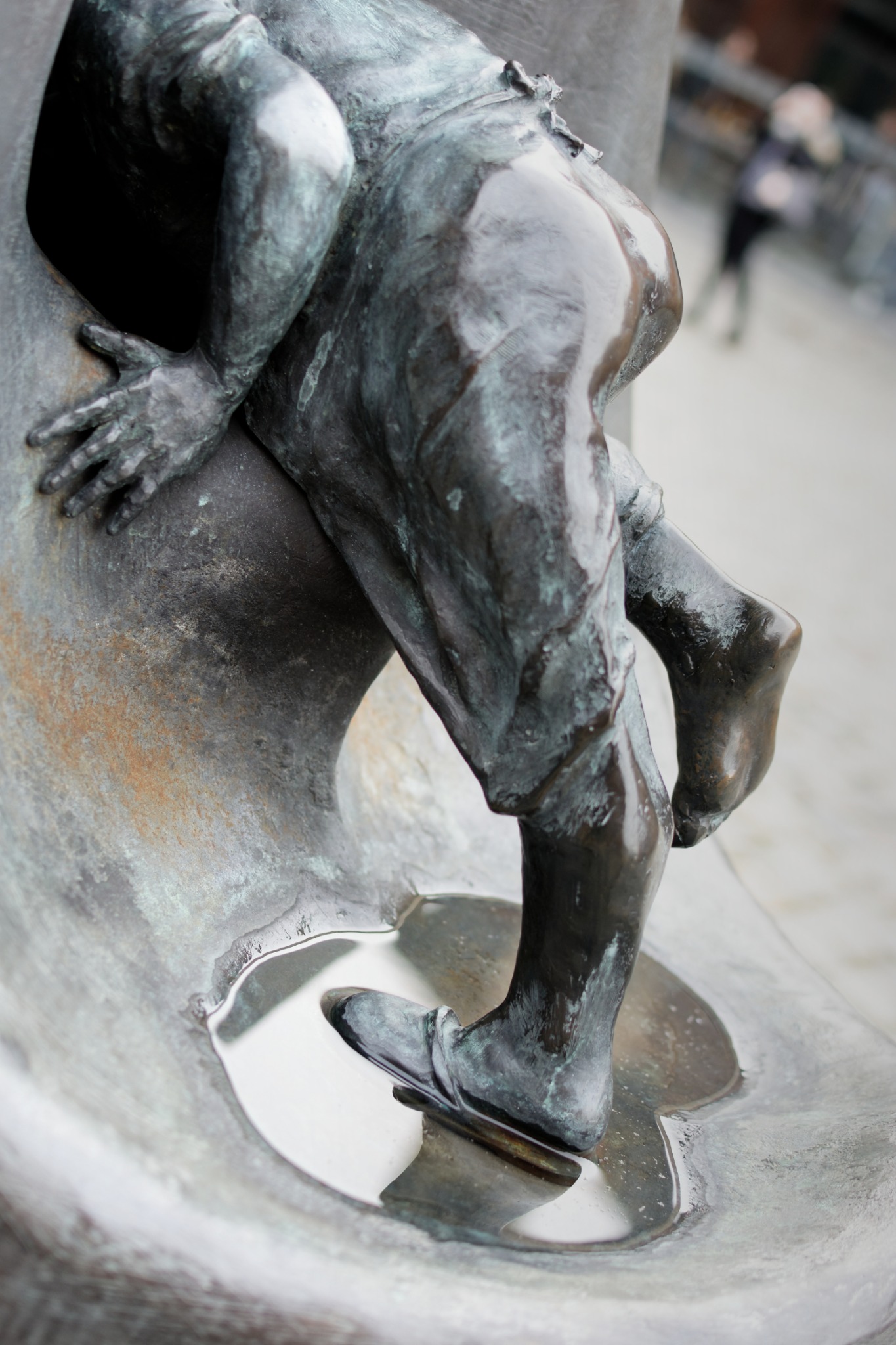 Brązowa rzeźba chłopca wspinającego się, z widocznymi detalami faktury i patyny. Woda zbiera się u podstawy, odbijając światło. Ujęcie z bliska, z rozmytym tłem.