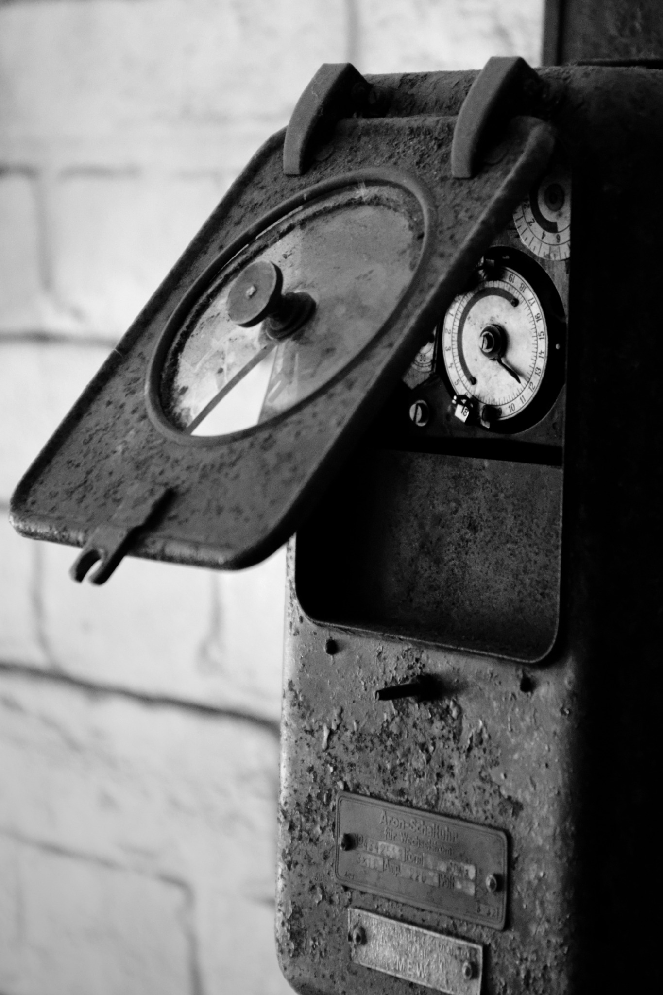 Czarno-białe zdjęcie starego, zardzewiałego licznika elektrycznego z otwartą klapką, ukazującego tarczę zegarową i tabliczki znamionowe. Detaliczny widok na urządzenie.
