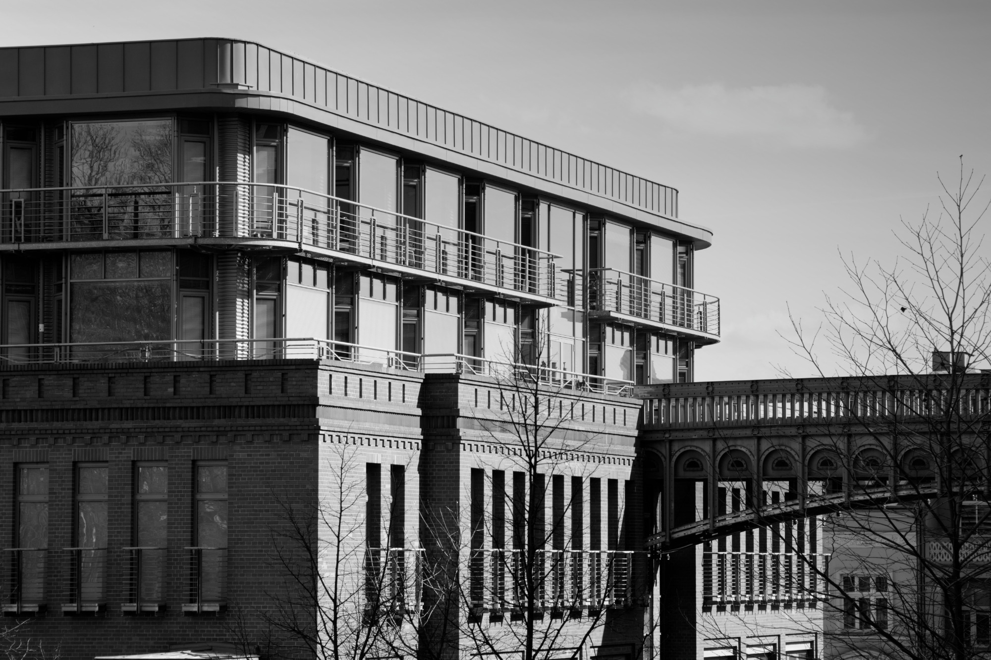 Czarno-białe zdjęcie budynku biurowego z balkonami i łącznikiem, widoczne nagie gałęzie drzew, architektura industrialna z elementami nowoczesnymi.