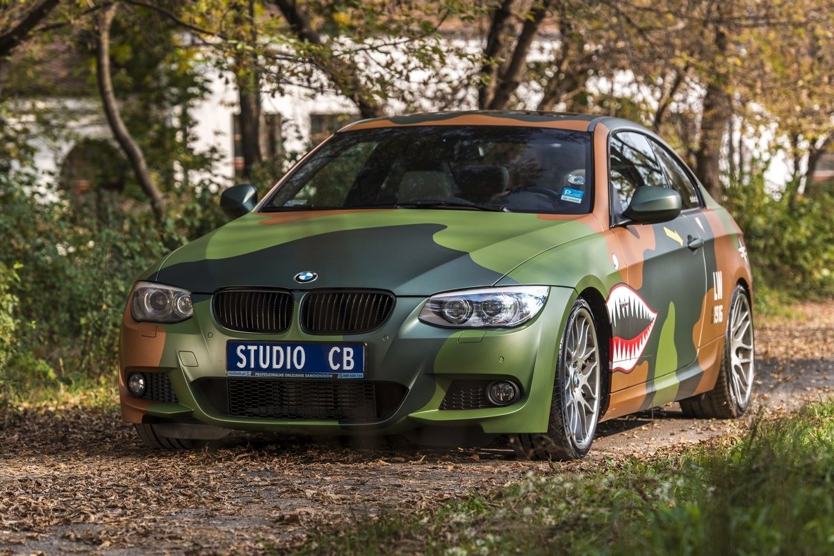 BMW E92 w matowym kamuflażu wojskowym z motywem rekina, zaparkowane na leśnej drodze. Tablica rejestracyjna 'STUDIO CB'.