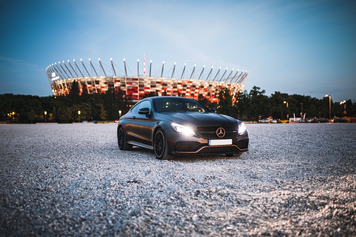 Matowy, czarny Mercedes C63 AMG Coupe na żwirowym parkingu, z włączonymi światłami, na tle Stadionu Narodowego o zmierzchu. Ujęcie z dołu, podkreślające sportowy charakter auta.