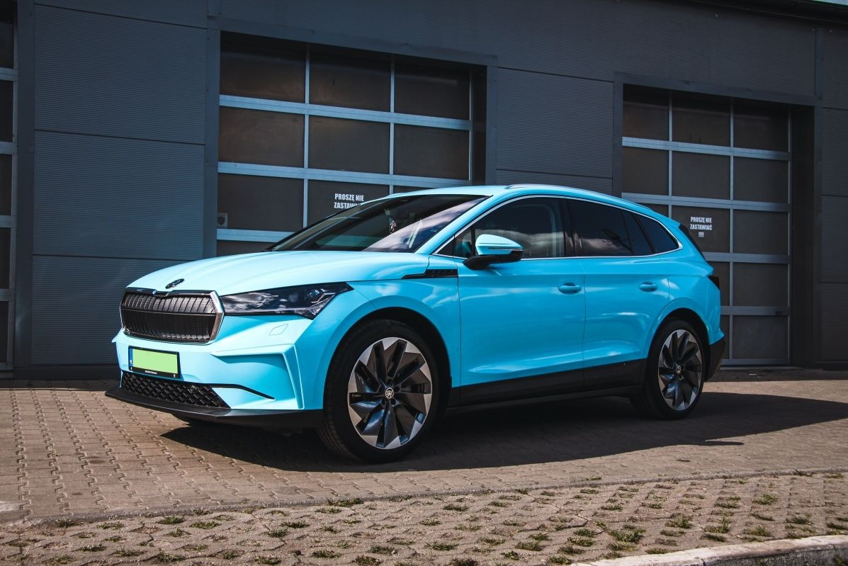 Błękitna Skoda Enyaq iV po detailingu, zaparkowana przed szarym budynkiem z bramami garażowymi. Widoczny połysk lakieru i czyste felgi. Ujęcie z boku, lekko od przodu.