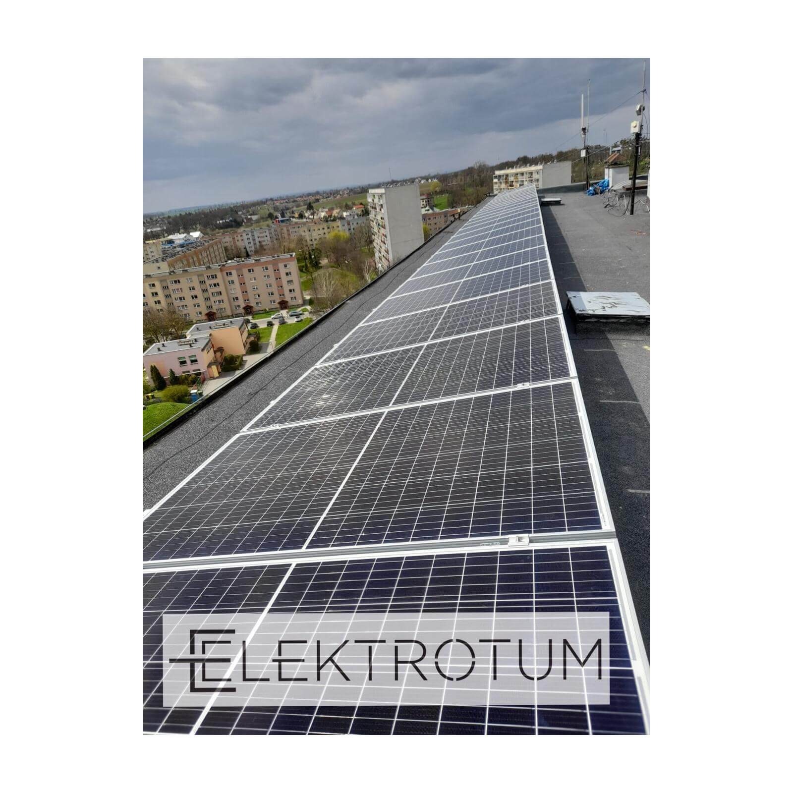 Widok z góry na rząd paneli fotowoltaicznych na dachu budynku, z widoczną panoramą miasta w tle i logo firmy na pierwszym planie.