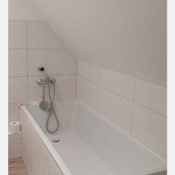 Małgorzata Kargol - Biała wanna obudowana płytkami w jasnej łazience na poddaszu. Bateria wannowa zamontowana na ścianie. Widoczne płytki na ścianach i podłodze.