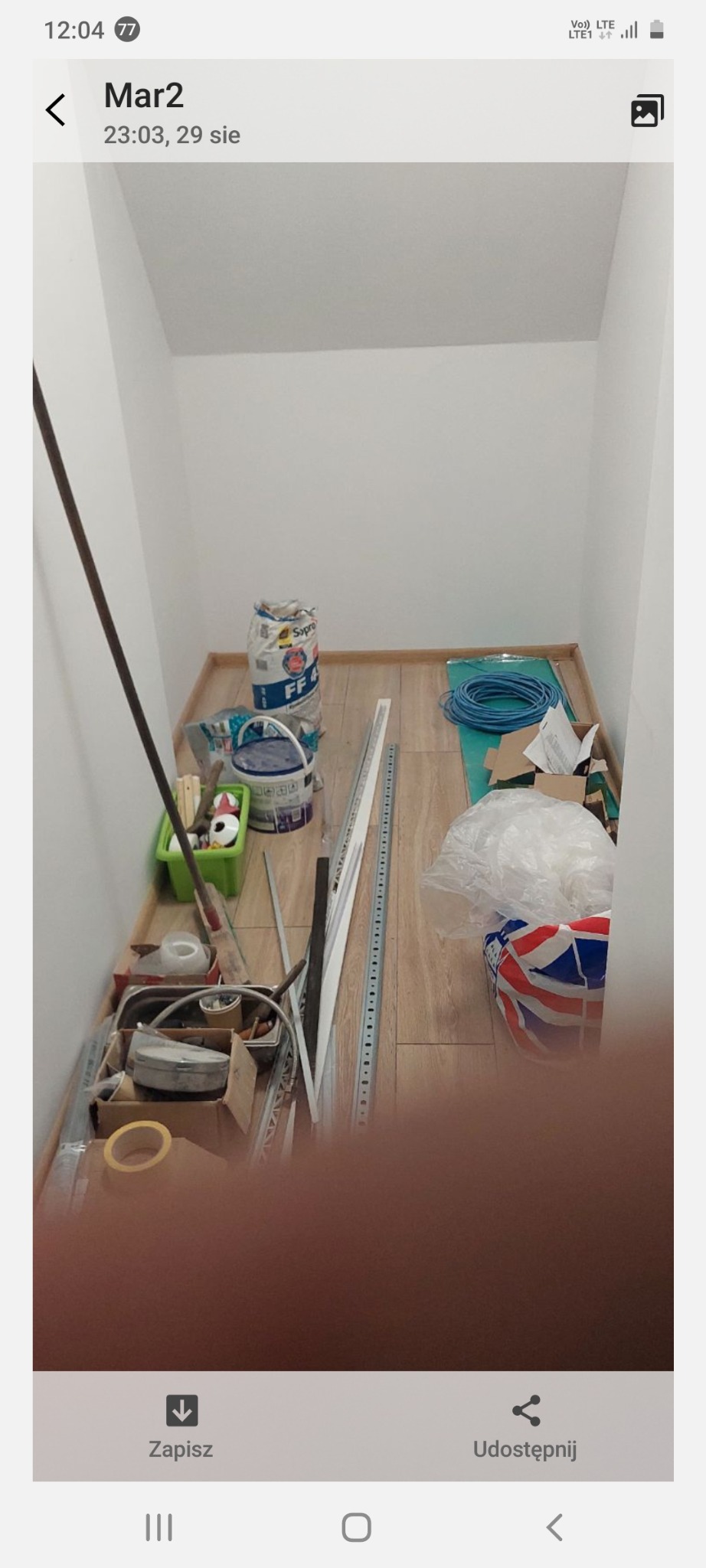 Kącik gospodarczy przed sprzątaniem: wiadro z farbą, worek z zaprawą, narzędzia i akcesoria malarskie w małym pomieszczeniu z białymi ścianami i drewnianą podłogą.