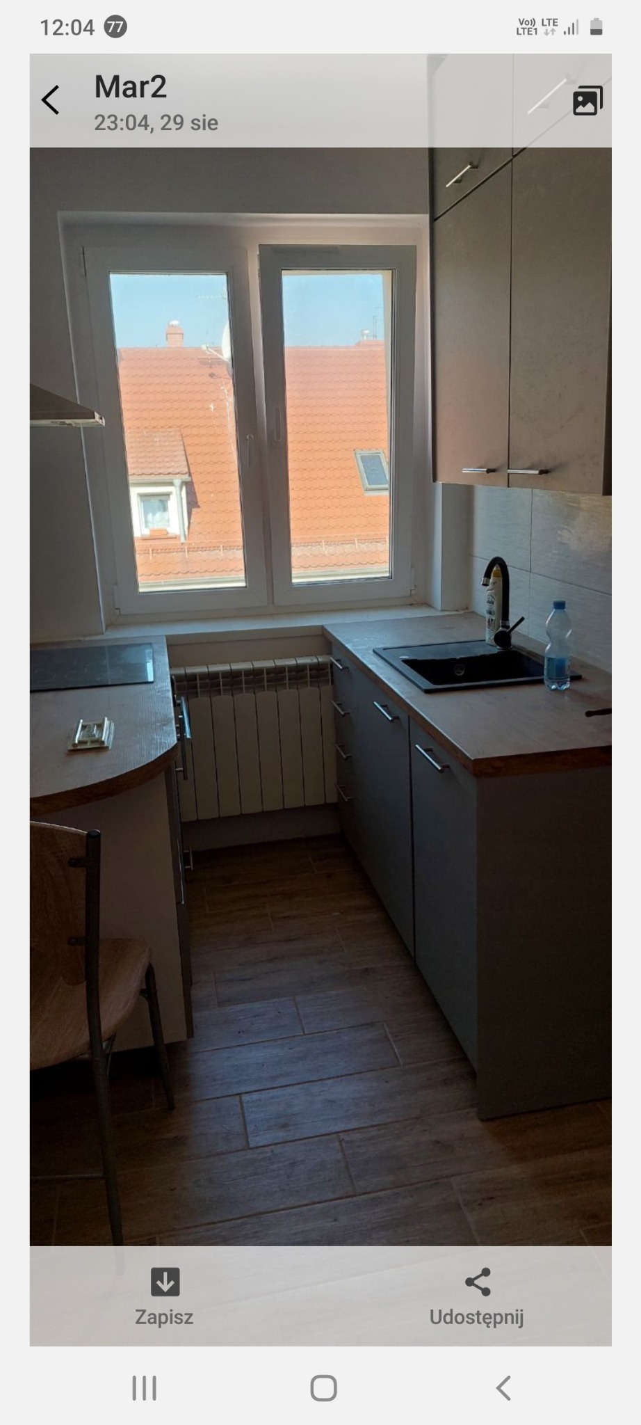 Jasna kuchnia z szarymi szafkami, czarnym zlewem i drewnianym blatem. Za oknem widok na czerwony dach. Na blacie butelka wody. Pod oknem grzejnik.