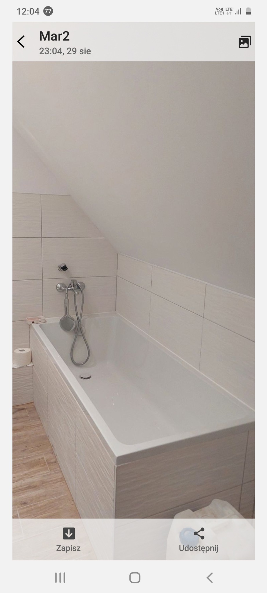 Biała wanna obudowana płytkami w jasnej łazience na poddaszu. Bateria wannowa zamontowana na ścianie. Widoczne płytki na ścianach i podłodze.