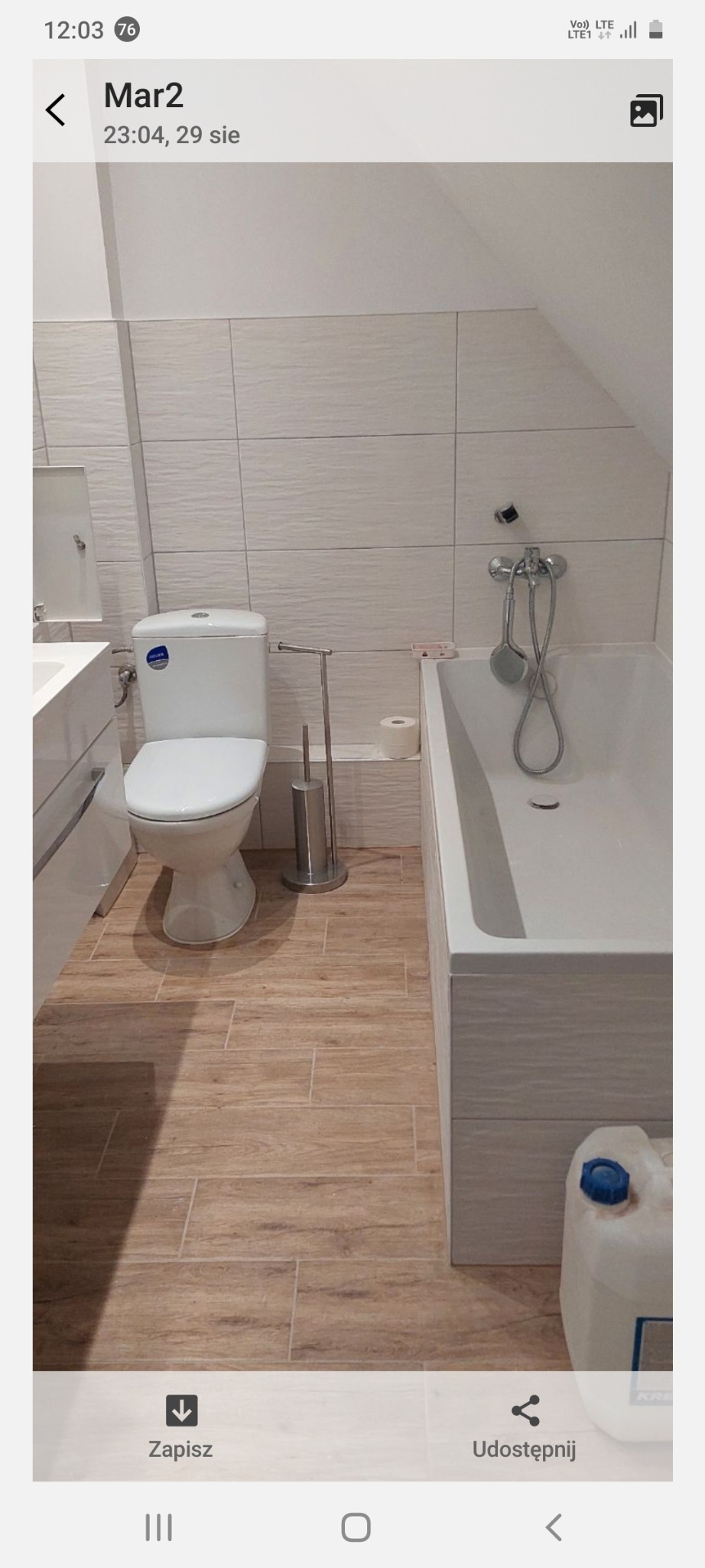 Jasna łazienka na poddaszu z beżowymi płytkami podłogowymi imitującymi drewno, białą armaturą i pojemnikiem z płynem do mycia pod wanną. Umywalka z szafką.