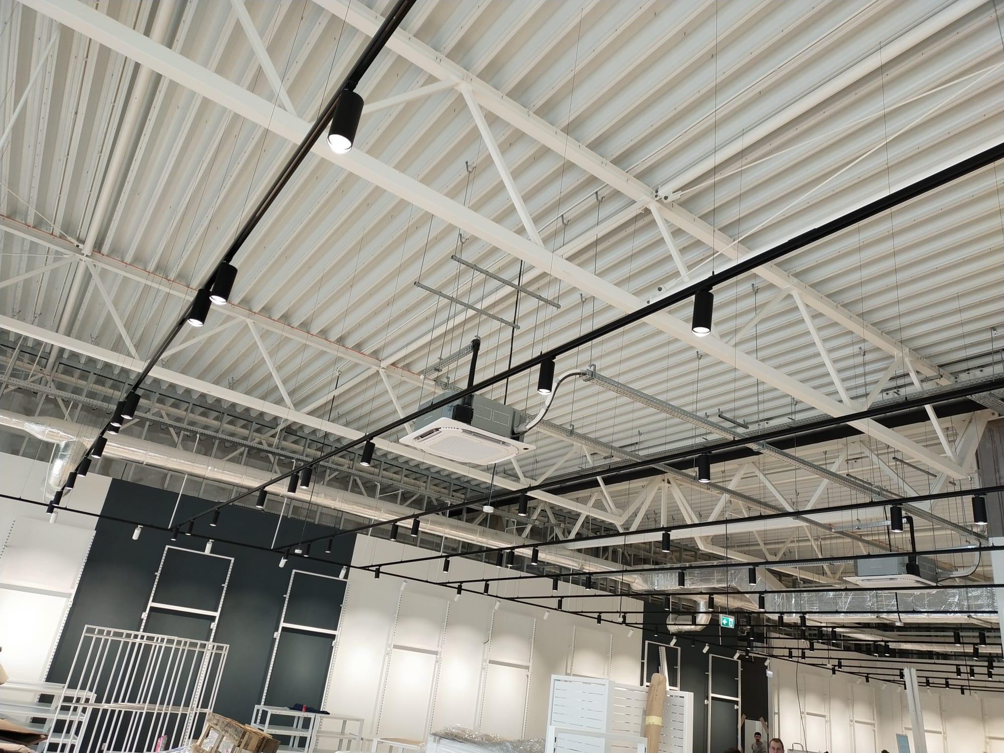 Nowoczesne oświetlenie szynowe z czarnymi reflektorami w dużej hali z białą, metalową konstrukcją sufitu. Widoczne elementy wentylacji i instalacji.