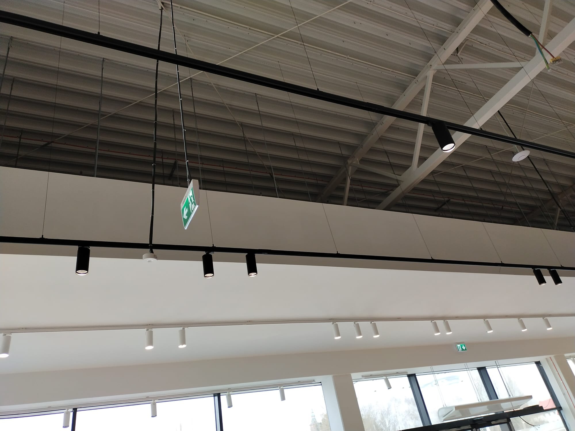 Biały sufit z zamontowanymi czarnymi reflektorami szynowymi i białymi punktowymi lampami. Widoczny znak ewakuacyjny i konstrukcja dachu.