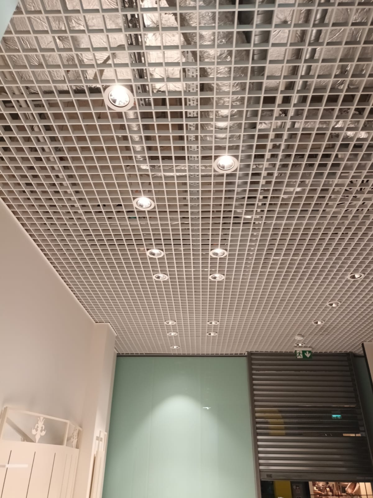 Biała, kratowa konstrukcja sufitowa z równomiernie rozmieszczonymi, okrągłymi lampami LED. Widoczny fragment ściany i roleta. Oświetlenie w przestrzeni komercyjnej.
