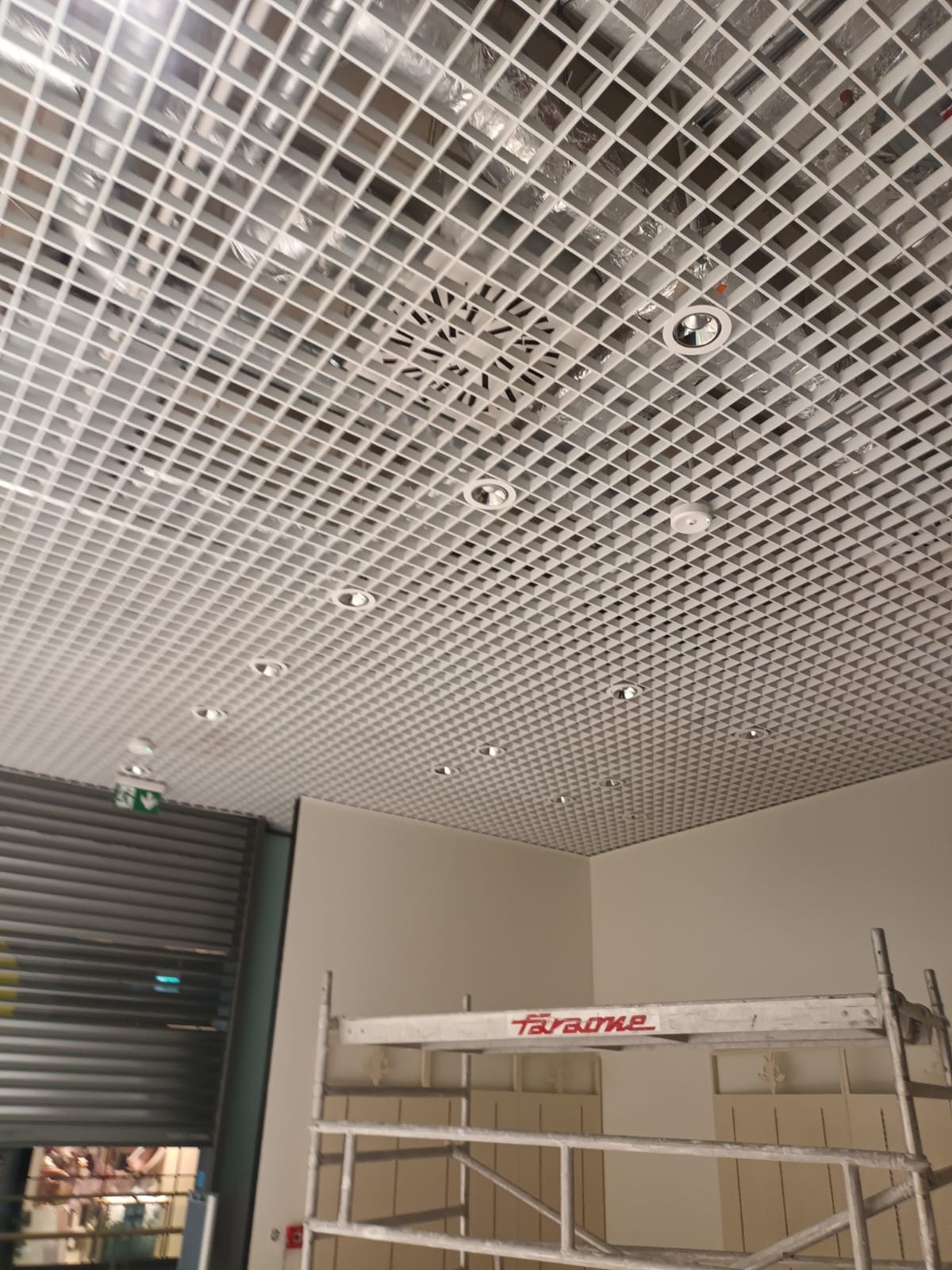 Kasetonowy sufit podwieszany z wbudowanymi punktami świetlnymi i kratką wentylacyjną. Widoczny fragment rusztowania i roleta.
