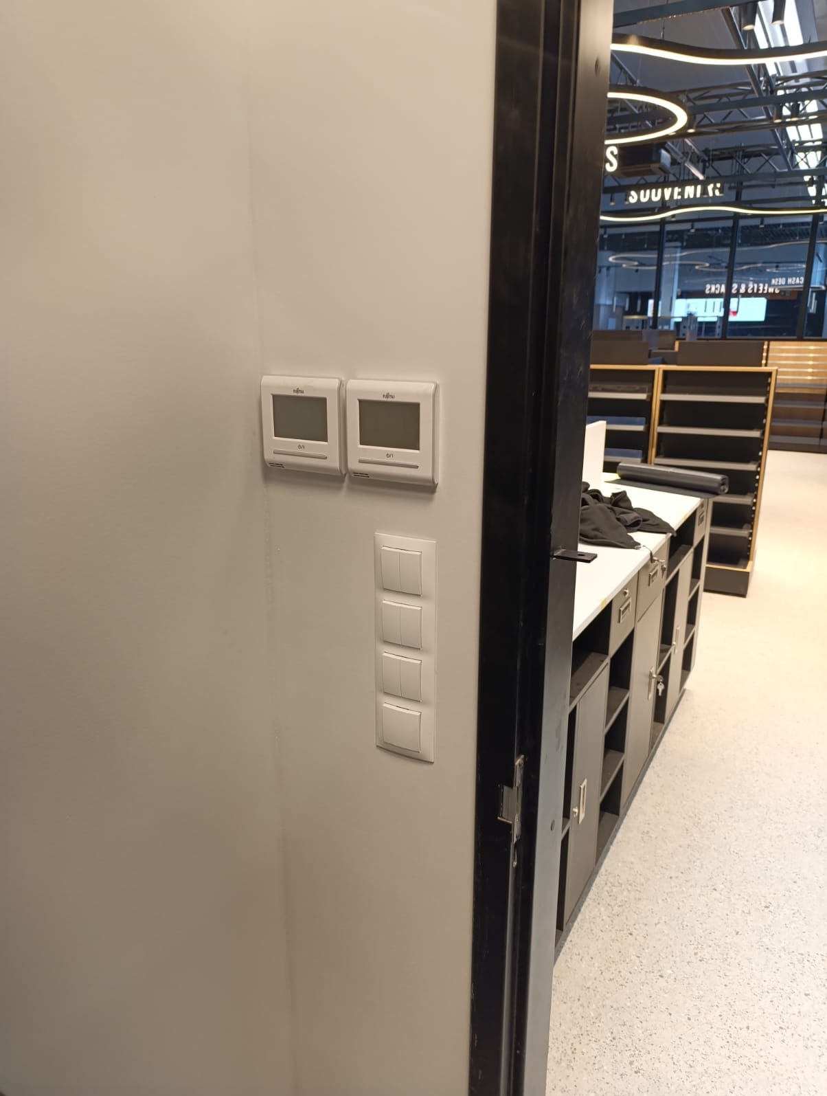 Dwa termostaty i poczwórny włącznik światła na ścianie w sklepie w Gdańsku. Widok zza futryny drzwi na wnętrze sklepu z regałami i oświetleniem.