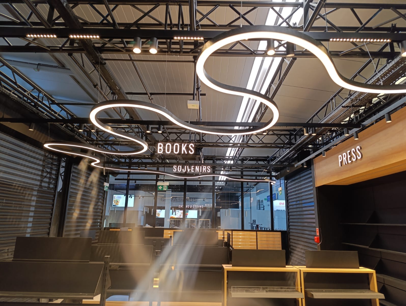 Nowoczesne oświetlenie LED w sklepie: wiszące lampy w kształcie fal, podwieszane reflektory punktowe, widoczne kable i konstrukcja sufitu. Napisy BOOKS, SOUVENIRS, PRESS.