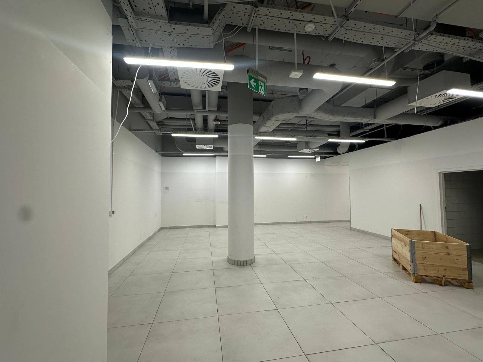 Wnętrze komercyjne przed pracami wykończeniowymi: widoczne instalacje pod sufitem, oświetlenie liniowe, wentylacja, kolumna nośna i pusta przestrzeń z paletą w rogu.