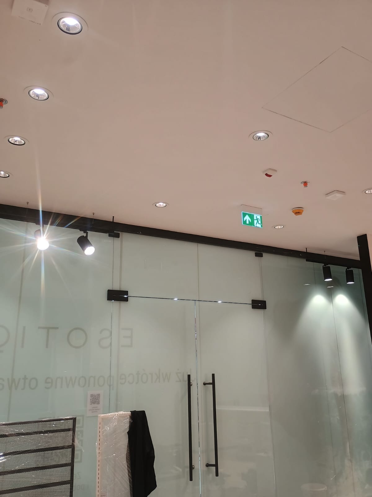 Sufit z zamontowanymi punktowymi lampami LED i reflektorami szynowymi, widoczny znak ewakuacyjny. Oświetlenie w nowoczesnym biurze lub sklepie.