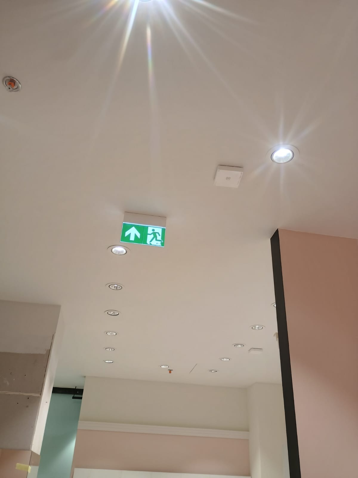 Biały sufit z zamontowanym oświetleniem punktowym i zielonym znakiem ewakuacyjnym. Widoczne elementy instalacji przeciwpożarowej i czujnik dymu.