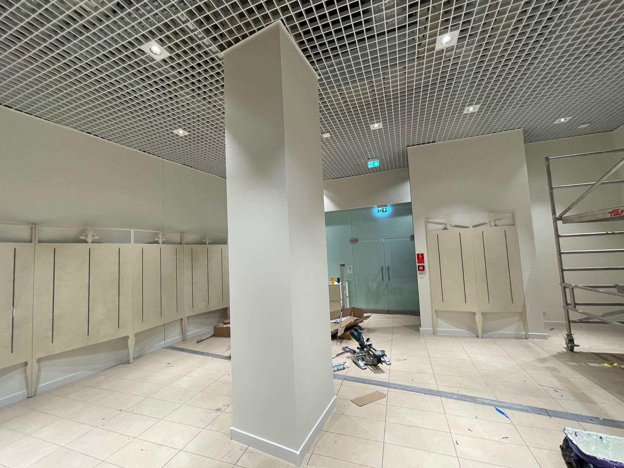 Wnętrze w trakcie prac wykończeniowych: kolumna, podwieszany sufit z oświetleniem, narzędzia i drabina. Widoczny etap prac przed ostatecznym wykończeniem.