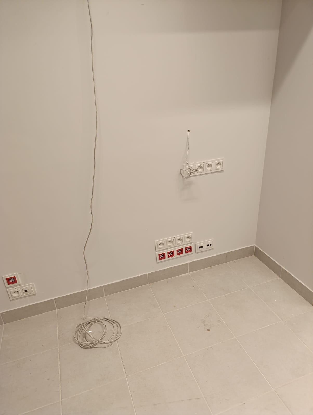 Białe ściany z zamontowanymi gniazdkami elektrycznymi i puszkami, zwisający kabel oraz zwinięty przewód na podłodze w nowym pomieszczeniu.