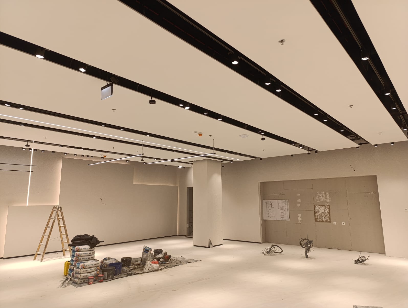 Nowoczesne oświetlenie sufitowe z czarnymi listwami i punktowymi lampami w przestronnym wnętrzu w trakcie wykańczania. Widoczna drabina i materiały budowlane.