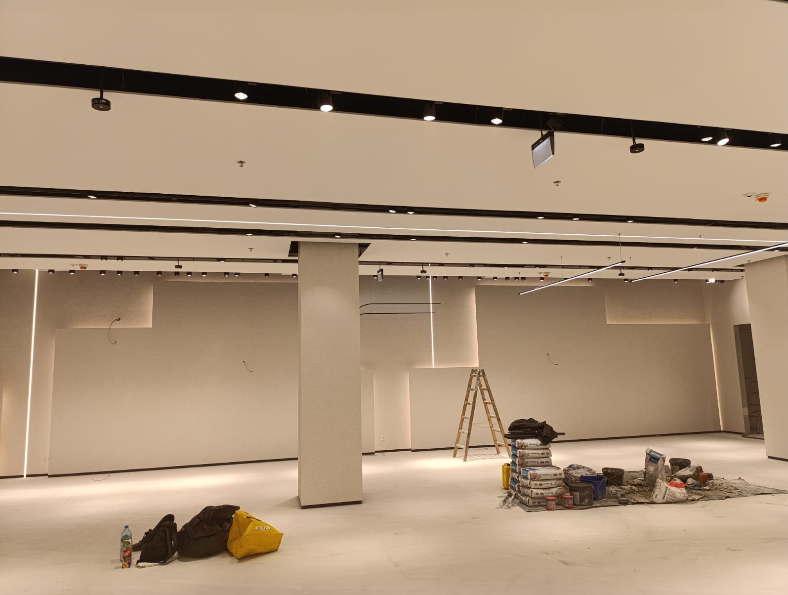 Wnętrze sklepu w trakcie wykańczania z zamontowanym oświetleniem liniowym i punktowym. Widoczne worki z zaprawą, drabina i narzędzia.