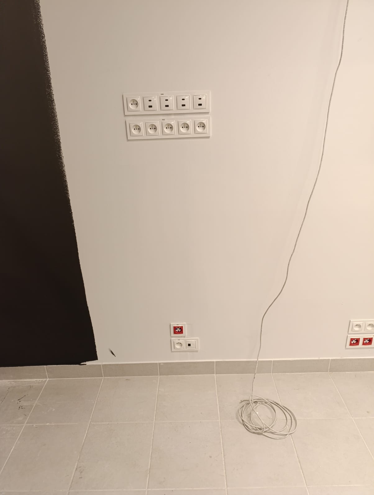 Biała ściana z zamontowanymi gniazdkami elektrycznymi i włącznikami, widoczne kable na podłodze i fragment pomalowanej na czarno ściany.