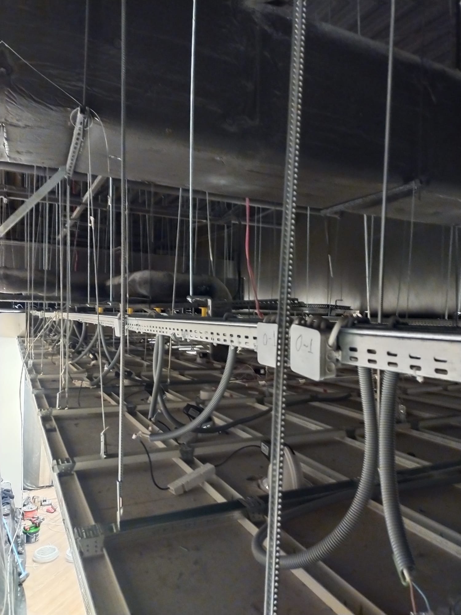 Sufit podwieszany z widocznymi elementami instalacji elektrycznej, w tym peszle i puszki z oznaczeniami, w trakcie prac montażowych. Widok z dołu.