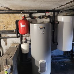 G3Service s.c. - Szary bojler Ferroli z podłączonymi rurami i zbiornikiem wyrównawczym w kotłowni o surowym wykończeniu. Widoczne elementy instalacji i narzędzia.