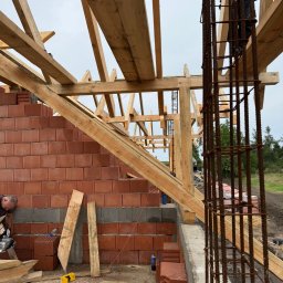 KonstrukcjeCNC - Wznoszenie konstrukcji dachu z drewnianych belek na murowanym budynku. Widoczny fragment zbrojenia i pracownik przy cięciu drewna.