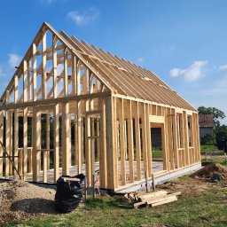 KonstrukcjeCNC - Surowa konstrukcja domu z drewna na fundamencie, widok z zewnątrz w słoneczny dzień. Na pierwszym planie sterta ziemi i czarny worek na śmieci.