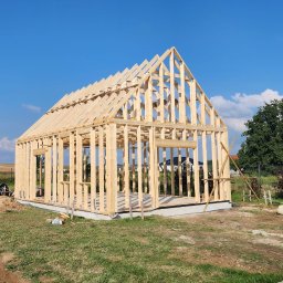 KonstrukcjeCNC - Konstrukcja szkieletowa domu jednorodzinnego w trakcie budowy, widok z zewnątrz, błękitne niebo w tle. Widoczne elementy drewniane i fundament.