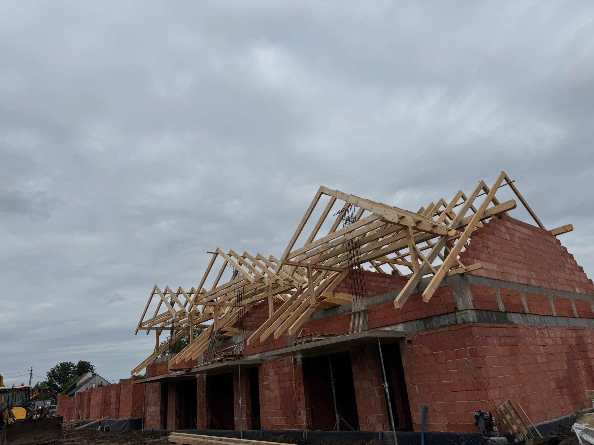 Wznoszenie konstrukcji dachu z drewnianych belek na budynku z czerwonej cegły pod pochmurnym niebem. Widoczny fragment koparki.