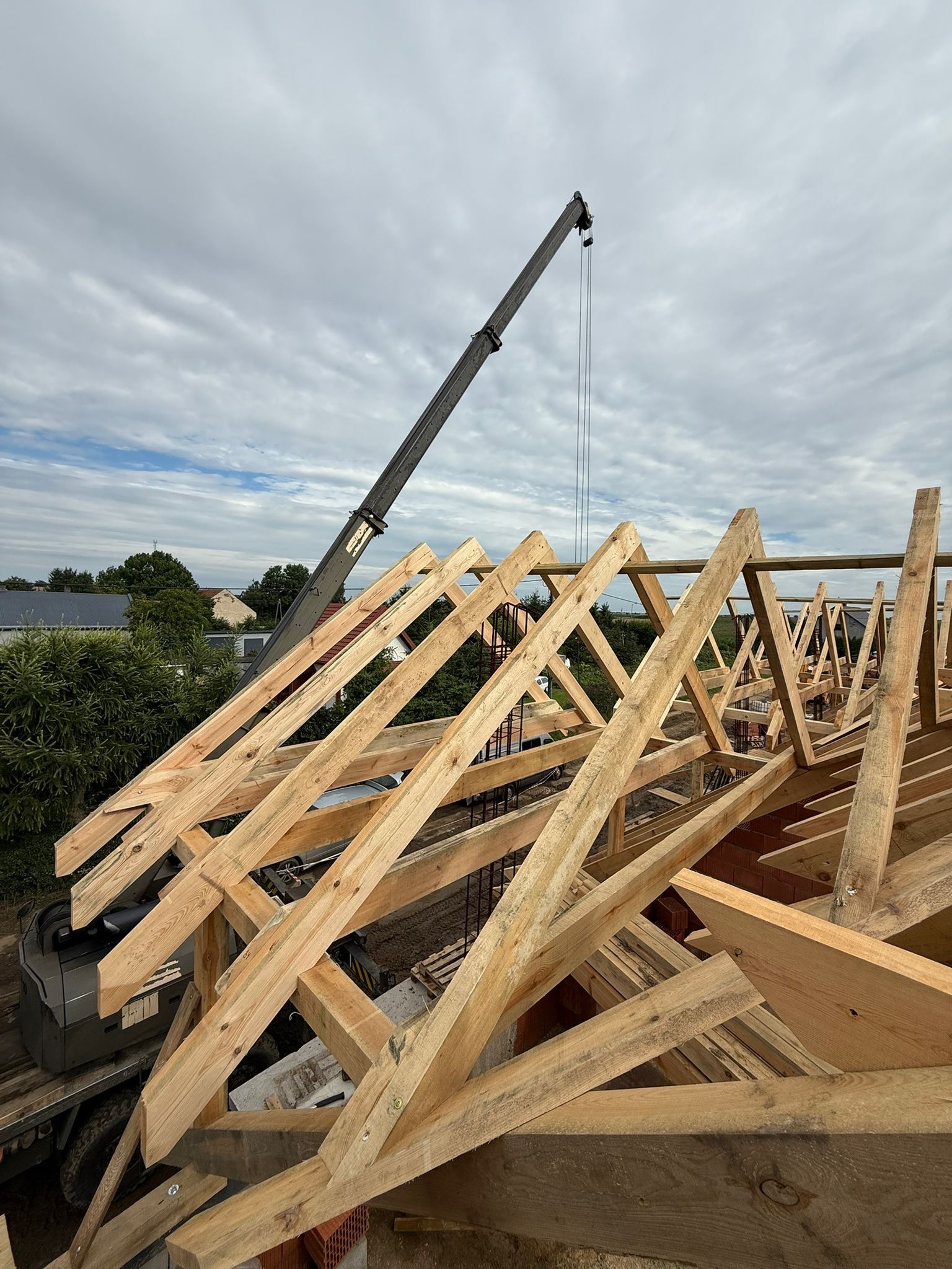 Wznoszenie drewnianej więźby dachowej z użyciem dźwigu na tle pochmurnego nieba. Widoczne elementy konstrukcyjne i fragmenty zabudowy mieszkalnej w tle.