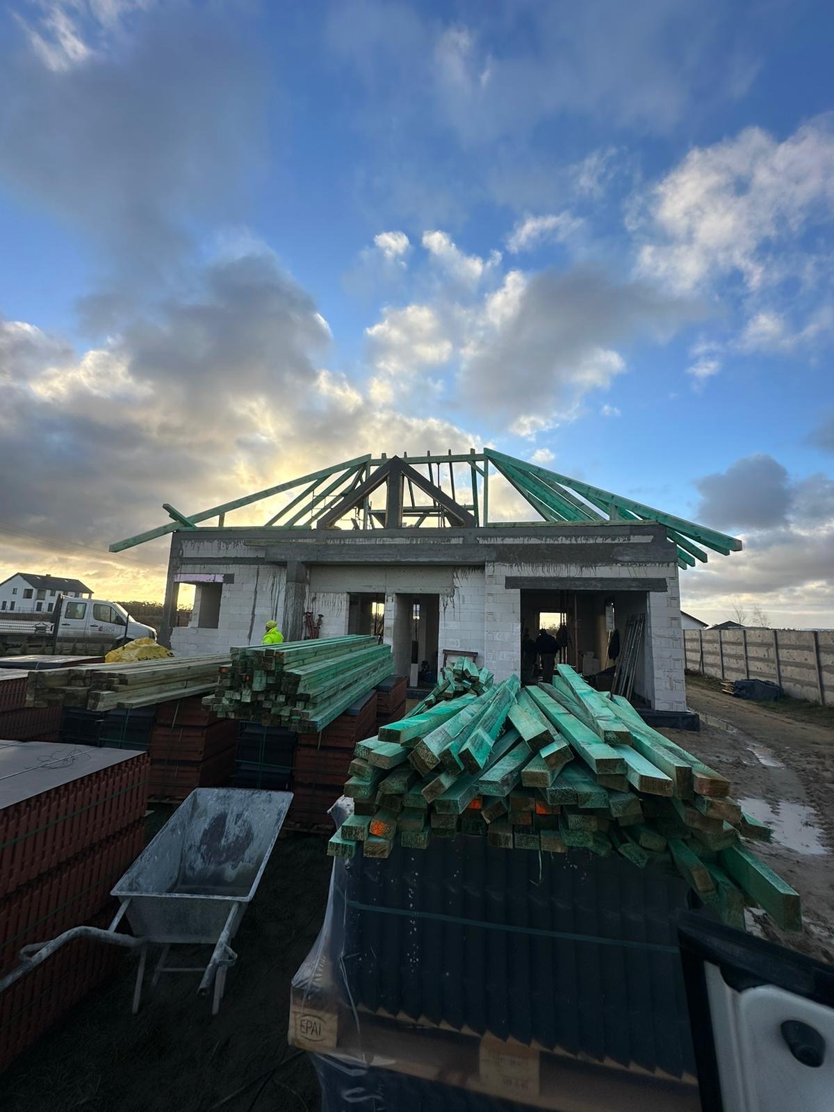 Budowa domu jednorodzinnego w trakcie realizacji: wzniesione mury z betonu komórkowego, częściowo wykonana więźba dachowa z zielonej impregnacji, tarcica budowlana na paletach.