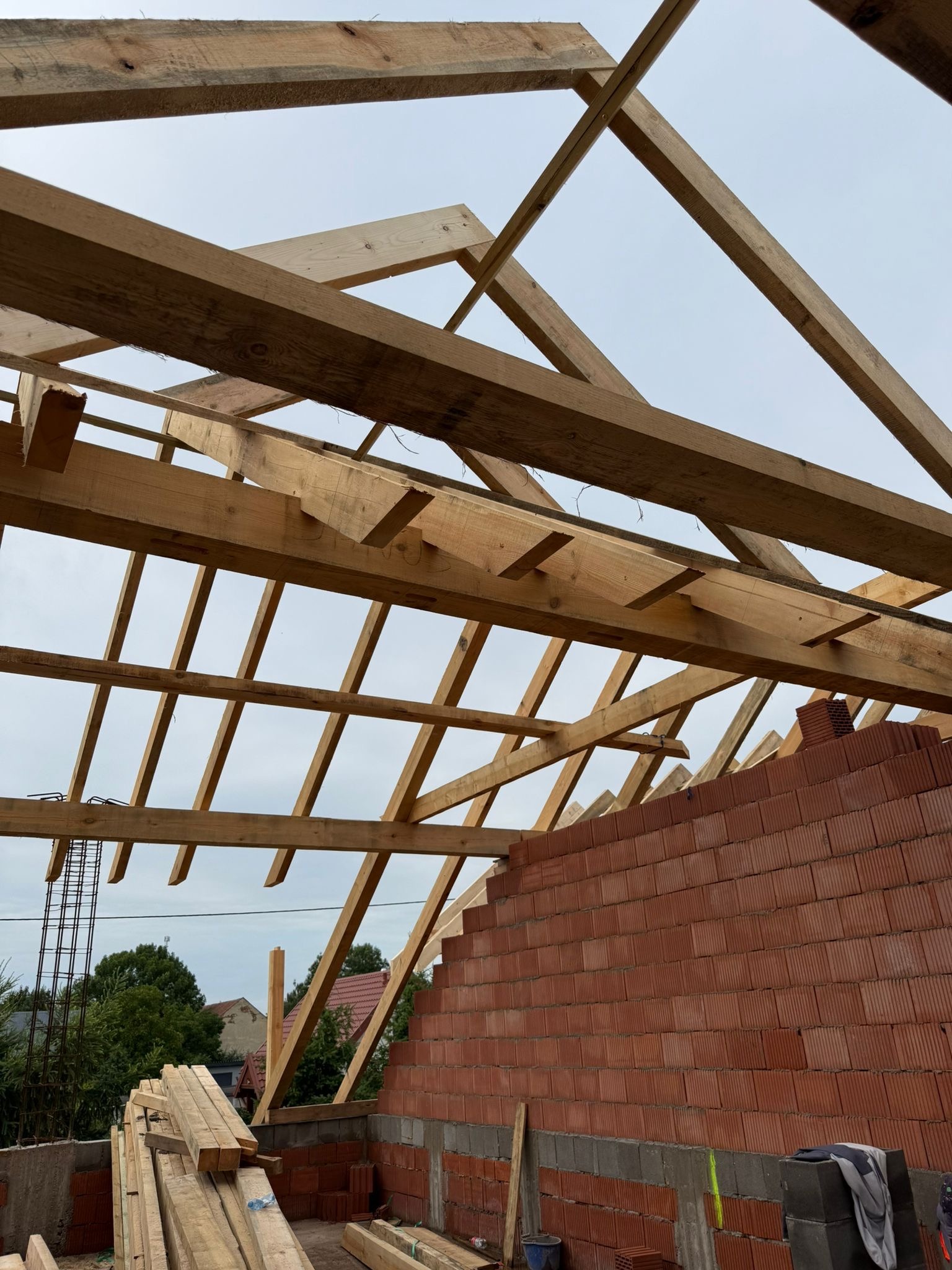 Wznoszenie drewnianej konstrukcji dachu z widoczną ścianą z czerwonej cegły dziurawki. Na pierwszym planie stos tarcicy. Widok z dołu na niebo.