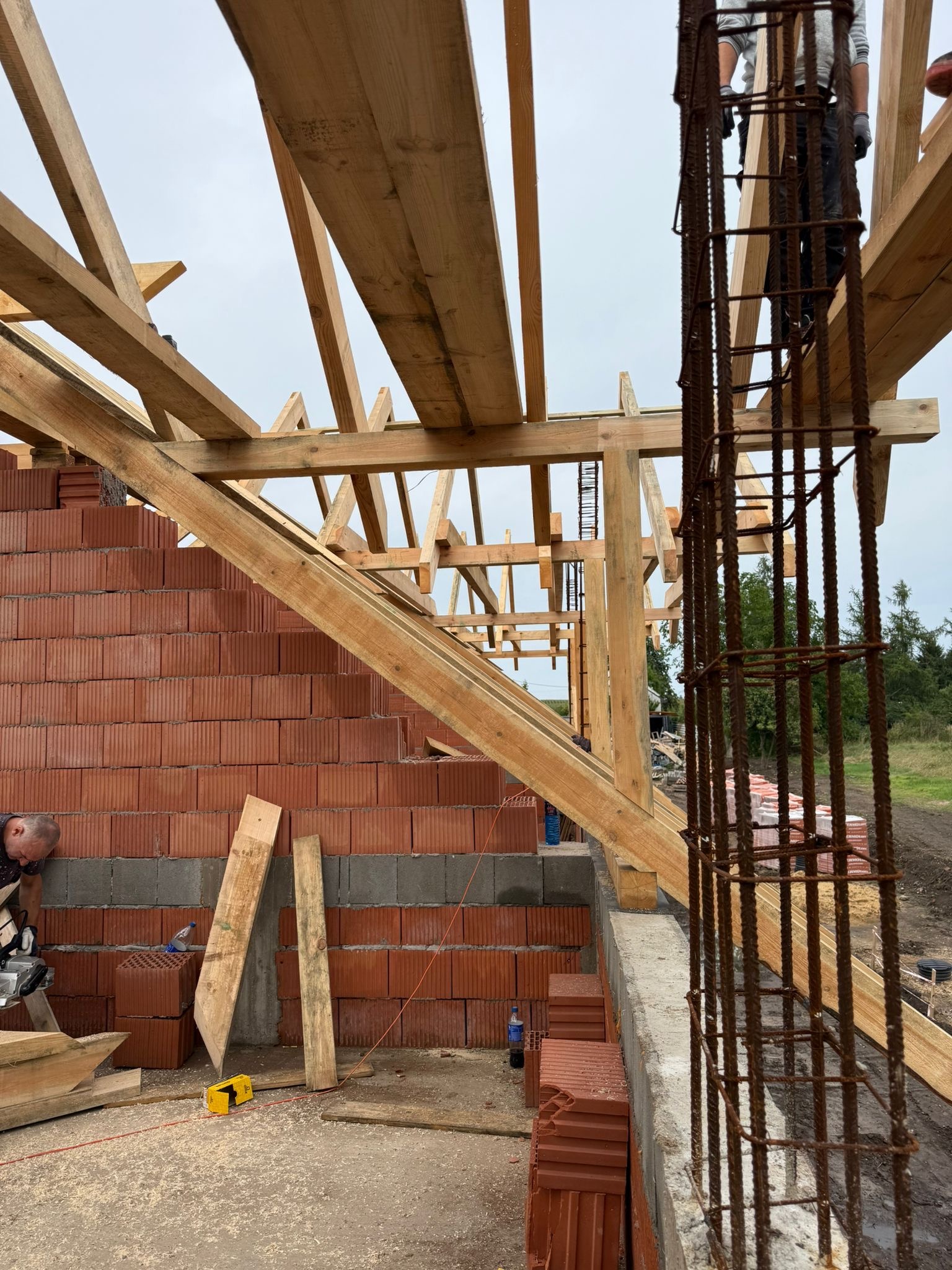 Wznoszenie konstrukcji dachu z drewnianych belek na murowanym budynku. Widoczny fragment zbrojenia i pracownik przy cięciu drewna.
