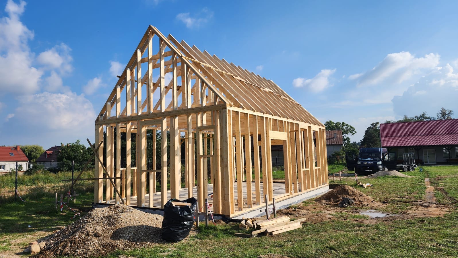 Wznoszenie drewnianej konstrukcji domu jednorodzinnego w technologii szkieletowej. Widoczne elementy więźby dachowej i ściany. W tle błękitne niebo i zabudowa.