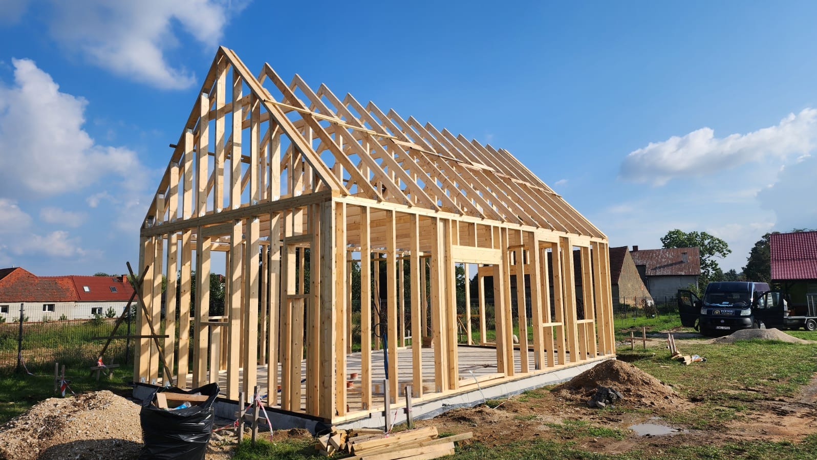 Konstrukcja szkieletowa domu w trakcie budowy, widok z zewnątrz w słoneczny dzień. Elementy drewniane tworzą ramy ścian i dachu na fundamencie.