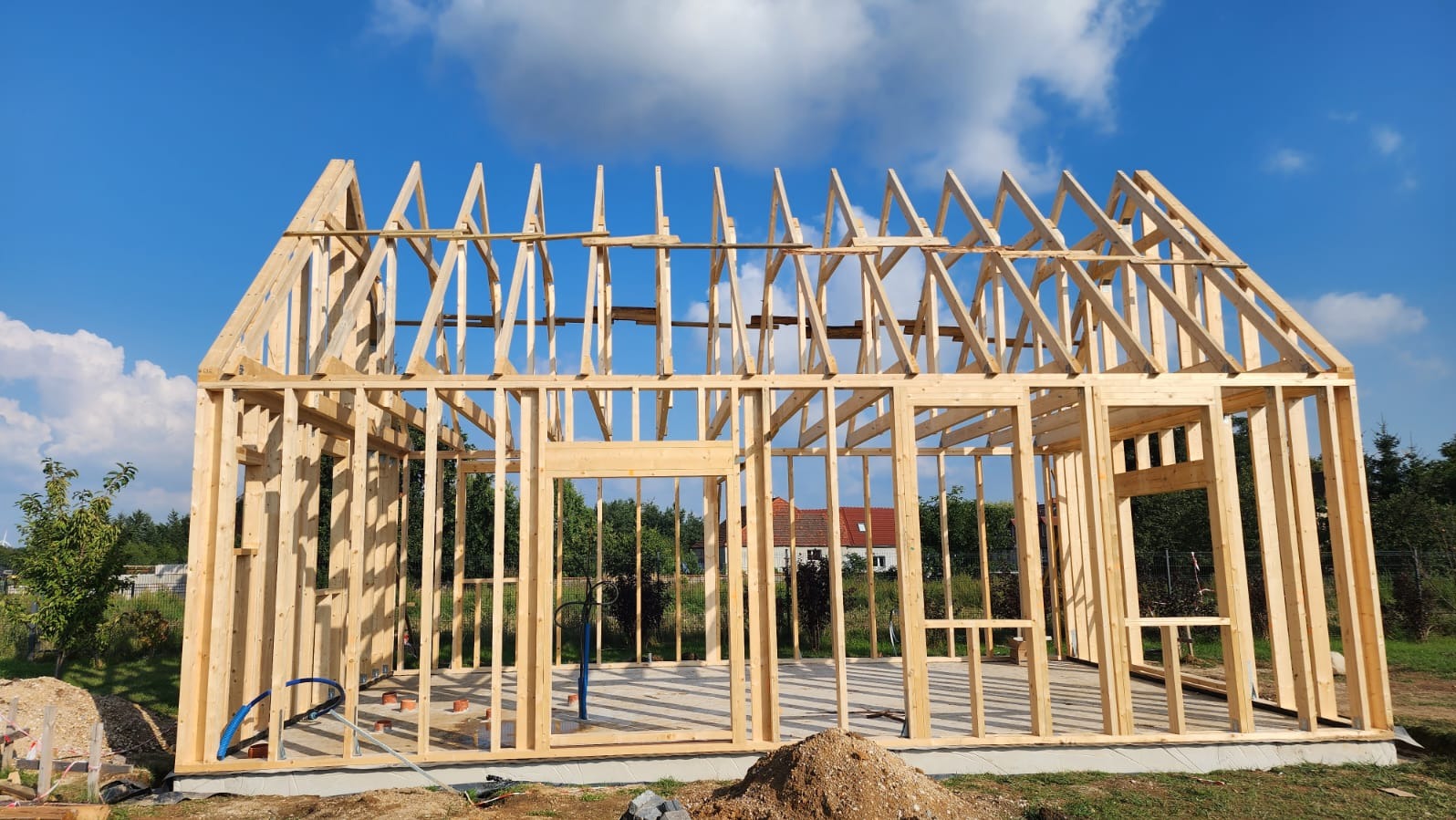 Konstrukcja szkieletowa domu w trakcie budowy, widoczne drewniane elementy ścian i dachu na tle błękitnego nieba. Na pierwszym planie ziemia i fragment instalacji.