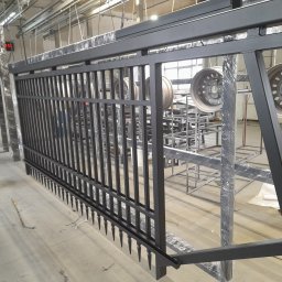 PAK PROTECT SPÓŁKA Z OGRANICZONĄ ODPOWIEDZIALNOŚCIĄ - Czarna, metalowa brama w trakcie malowania proszkowego w hali produkcyjnej. Widoczne elementy konstrukcyjne i zawieszone felgi samochodowe.