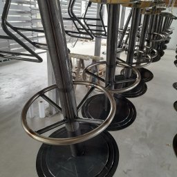 PAK PROTECT SPÓŁKA Z OGRANICZONĄ ODPOWIEDZIALNOŚCIĄ - Rząd metalowych krzeseł barowych w trakcie procesu malowania proszkowego, zawieszonych na hakach w hali produkcyjnej. Widoczne elementy konstrukcyjne i surowy wygląd.