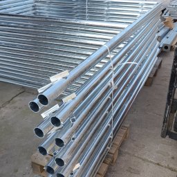 PAK PROTECT SPÓŁKA Z OGRANICZONĄ ODPOWIEDZIALNOŚCIĄ - Stos ocynkowanych, metalowych słupków ogrodzeniowych na palecie, przygotowanych do transportu. W tle widoczne panele ogrodzeniowe i wózek widłowy.