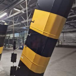 PAK PROTECT SPÓŁKA Z OGRANICZONĄ ODPOWIEDZIALNOŚCIĄ - Elementy konstrukcyjne w czarno-żółte pasy, zawieszone w hali produkcyjnej, po procesie malowania proszkowego. Widoczne detale mocowań i metalowa konstrukcja dachu.