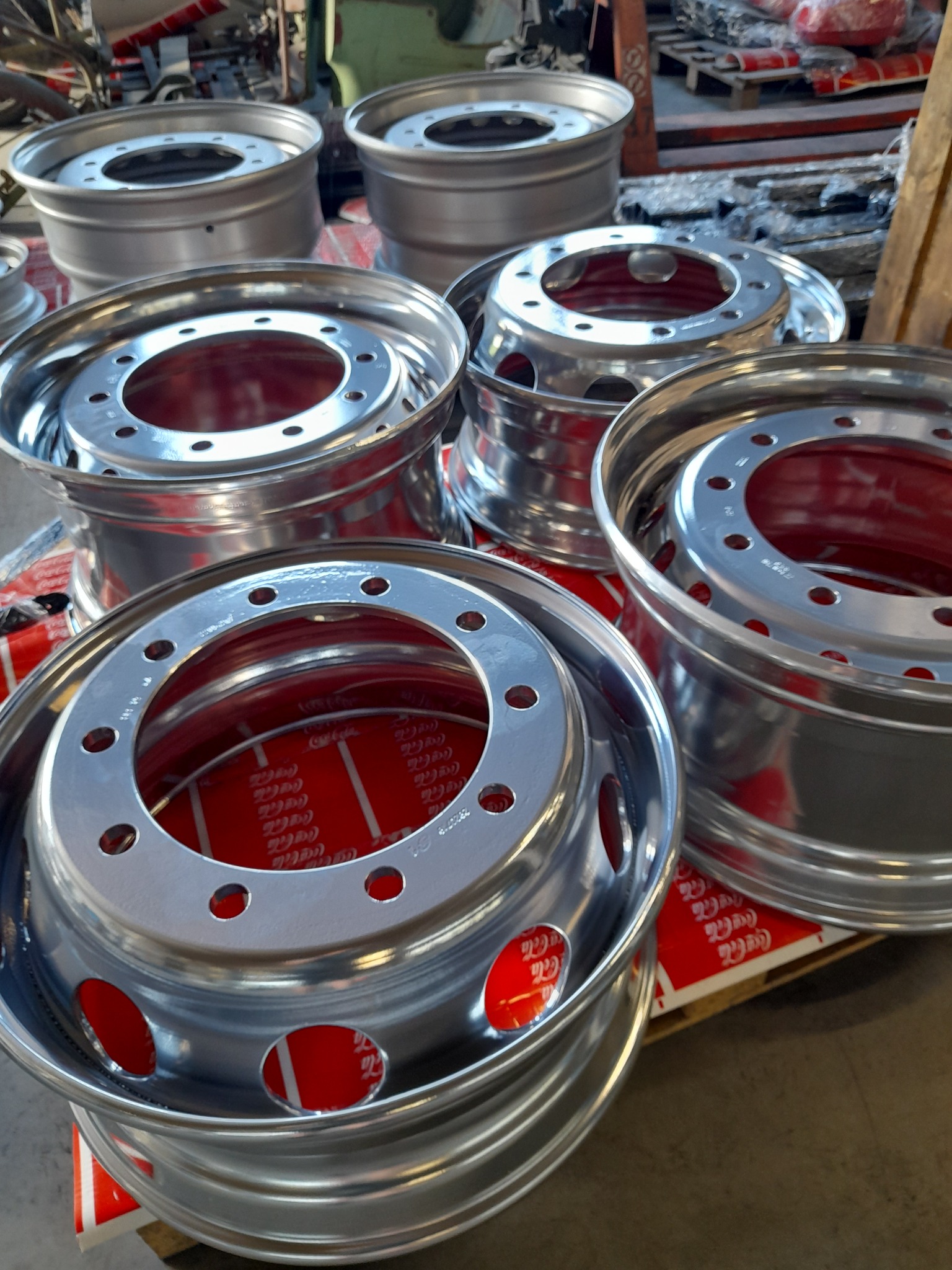 Błyszczące, srebrne felgi samochodowe z otworami na śruby, ułożone na palecie z czerwonym nadrukiem. Widoczne detale wykończenia i odbicia światła.