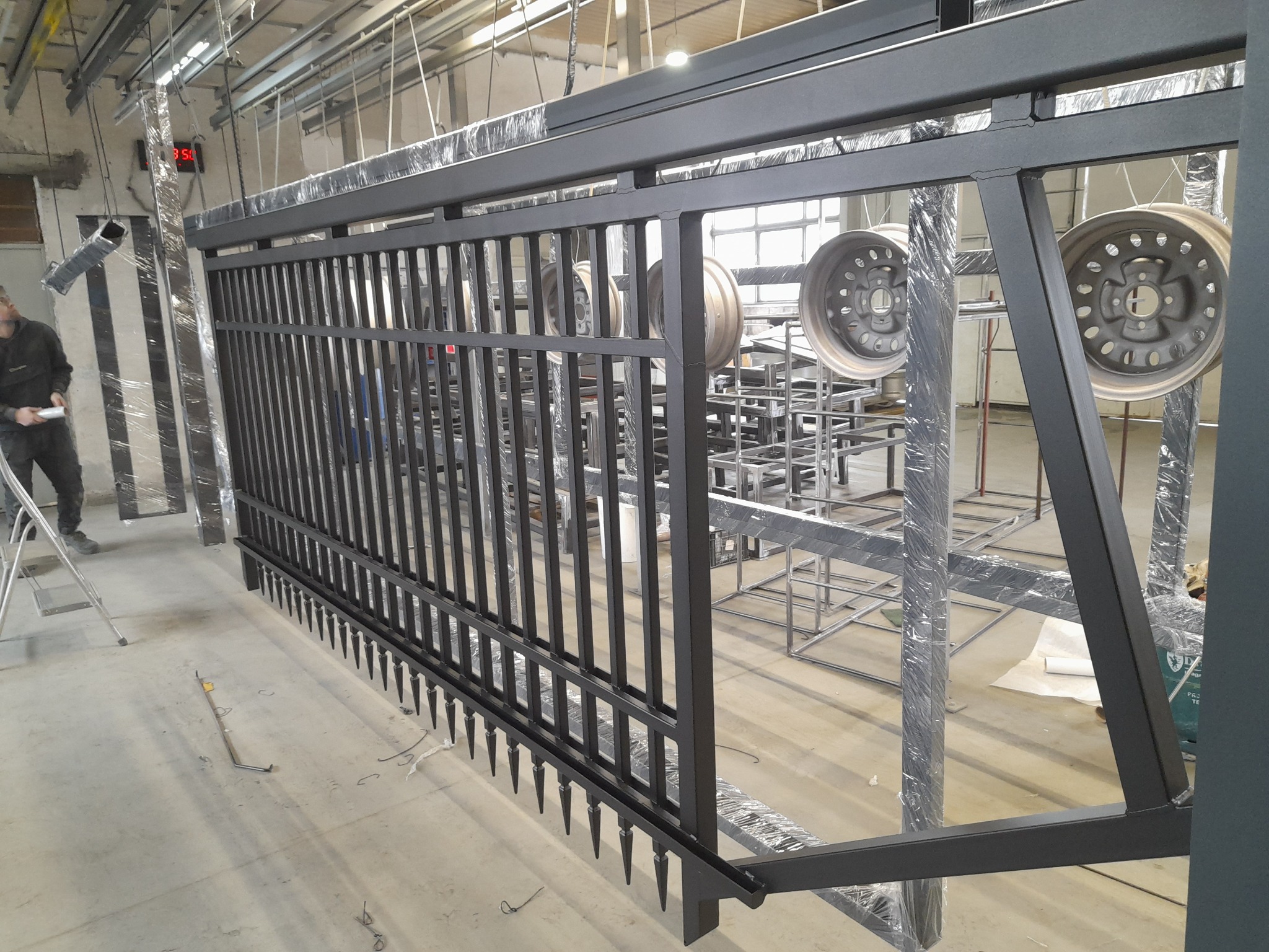 Czarna, metalowa brama w trakcie malowania proszkowego w hali produkcyjnej. Widoczne elementy konstrukcyjne i zawieszone felgi samochodowe.