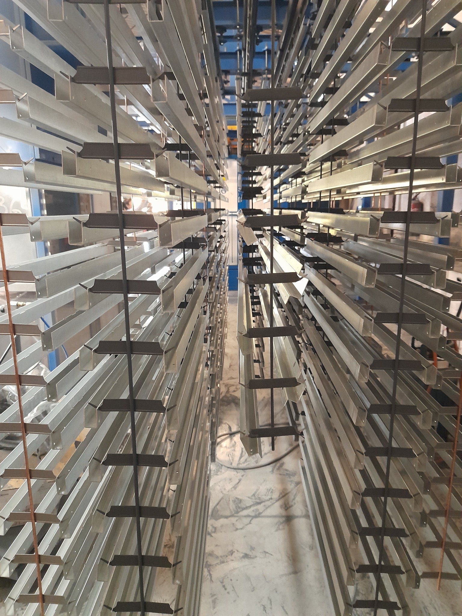 Metalowe elementy konstrukcyjne zawieszone na wieszakach, przygotowane do malowania proszkowego. Widok z dołu, ukazujący perspektywę i powtarzalność elementów.