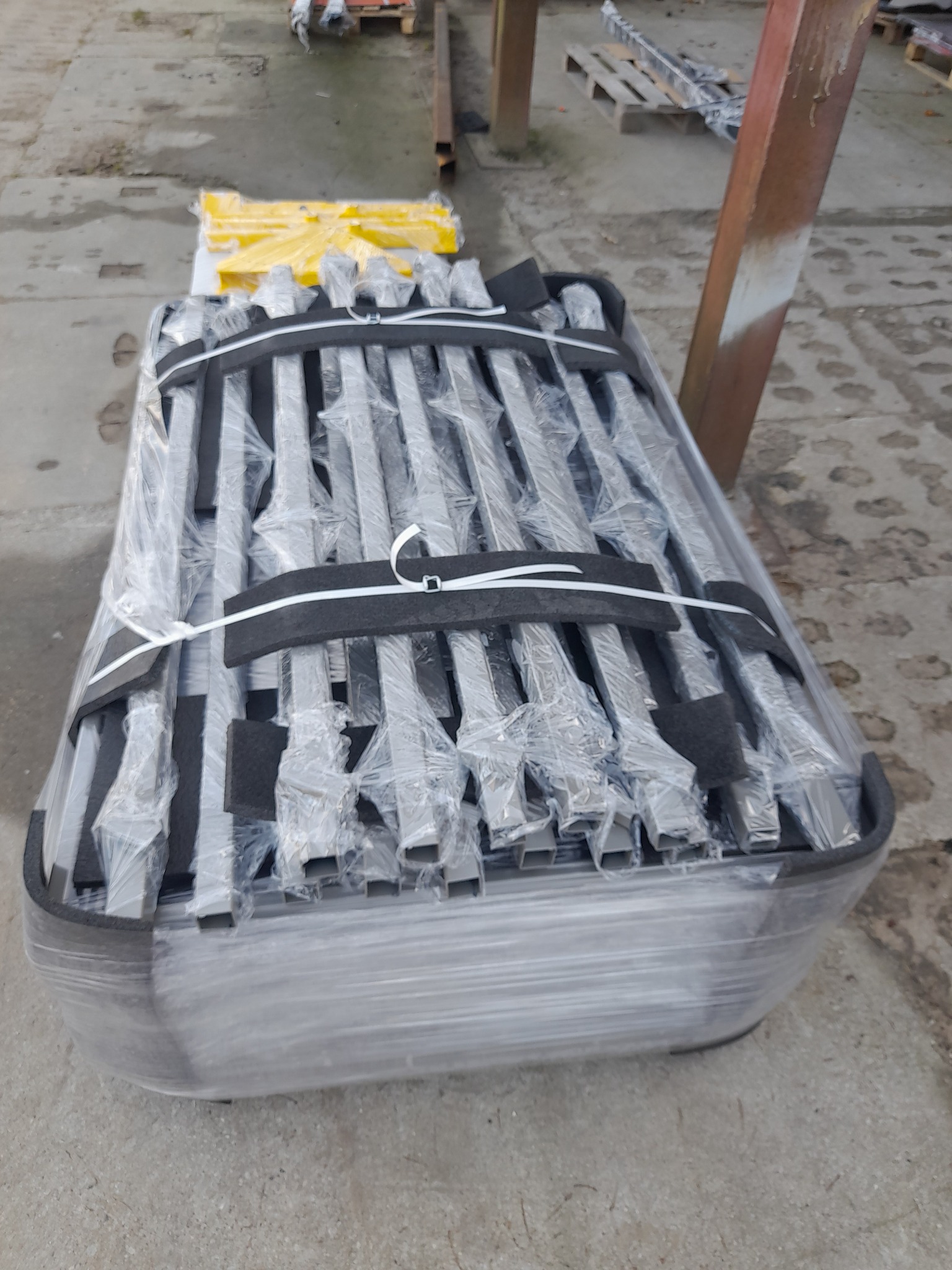 Zestaw metalowych słupków ogrodzeniowych, zabezpieczonych folią i spiętych taśmami, gotowych do transportu. Widoczne elementy montażowe i palety w tle.