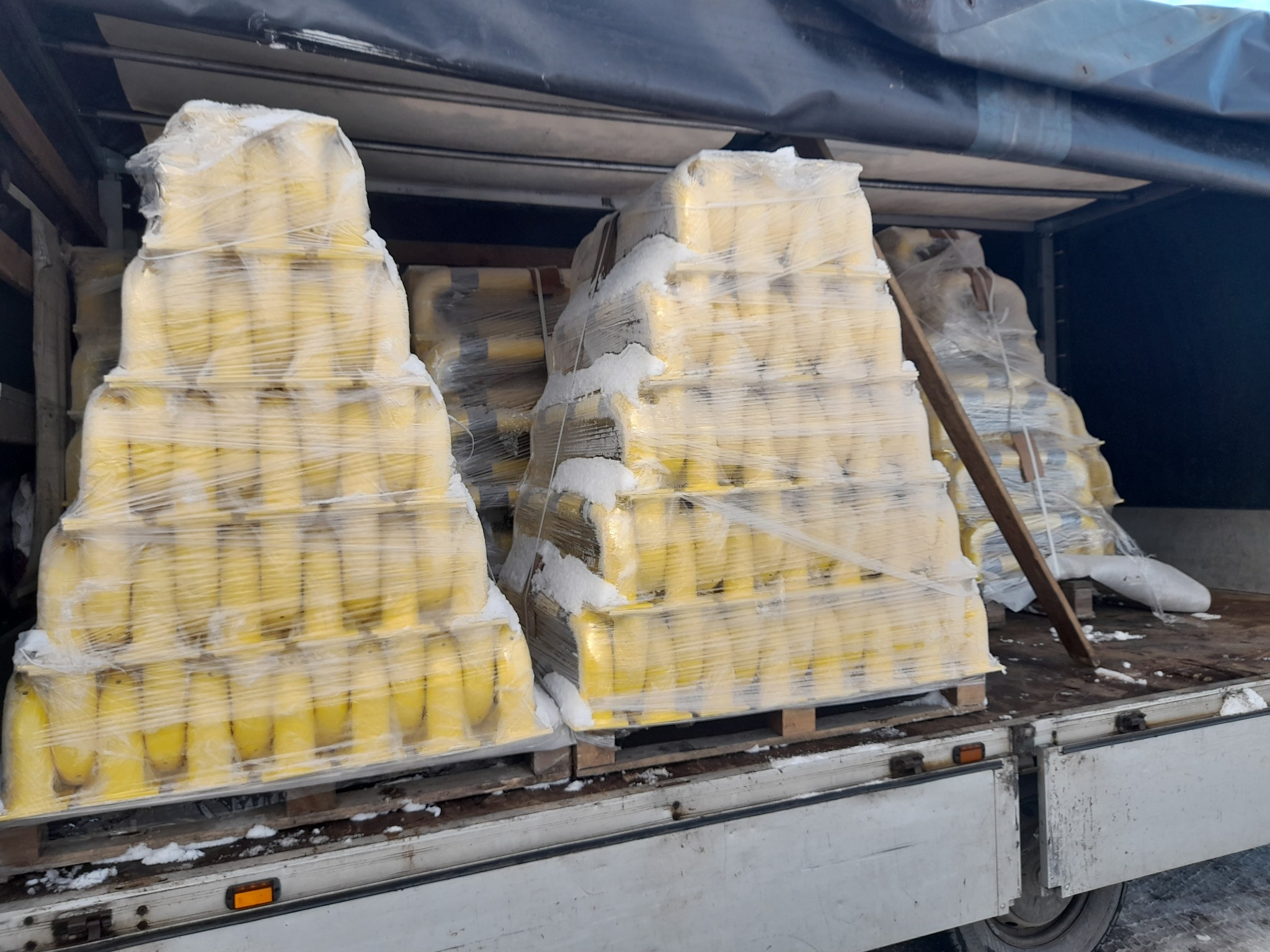 Żółte słupki ogrodzeniowe, spakowane na paletach i owinięte folią, przewożone w przestrzeni ładunkowej ciężarówki. Widoczne ślady śniegu.